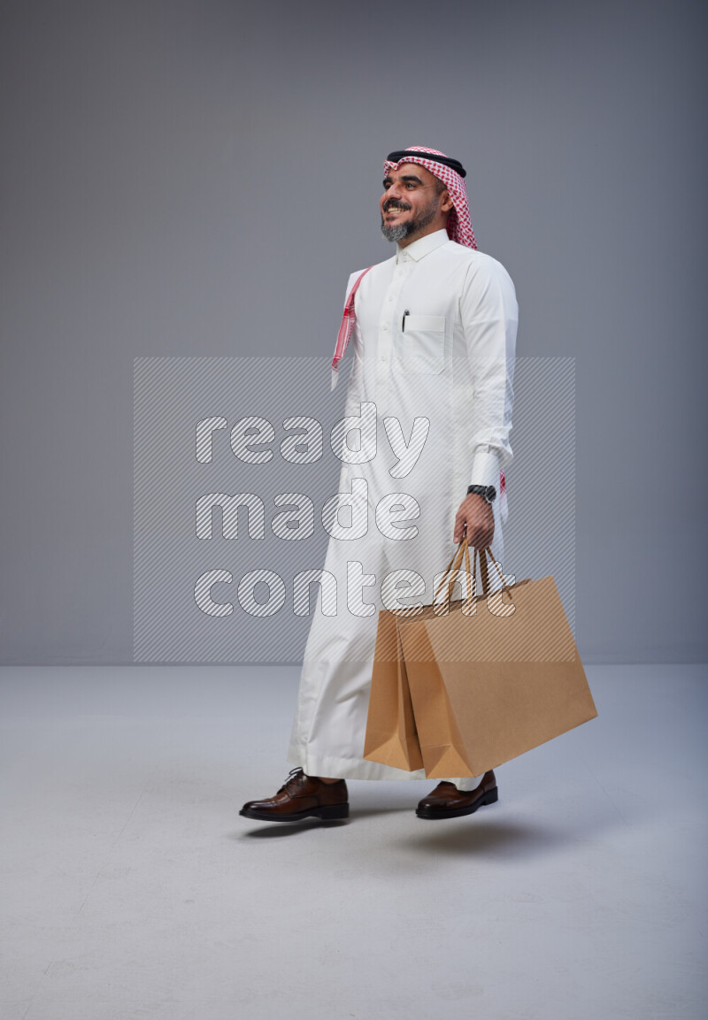 Saudi man Wearing Thob and red Shomag standing holding shopping bag on Gray background