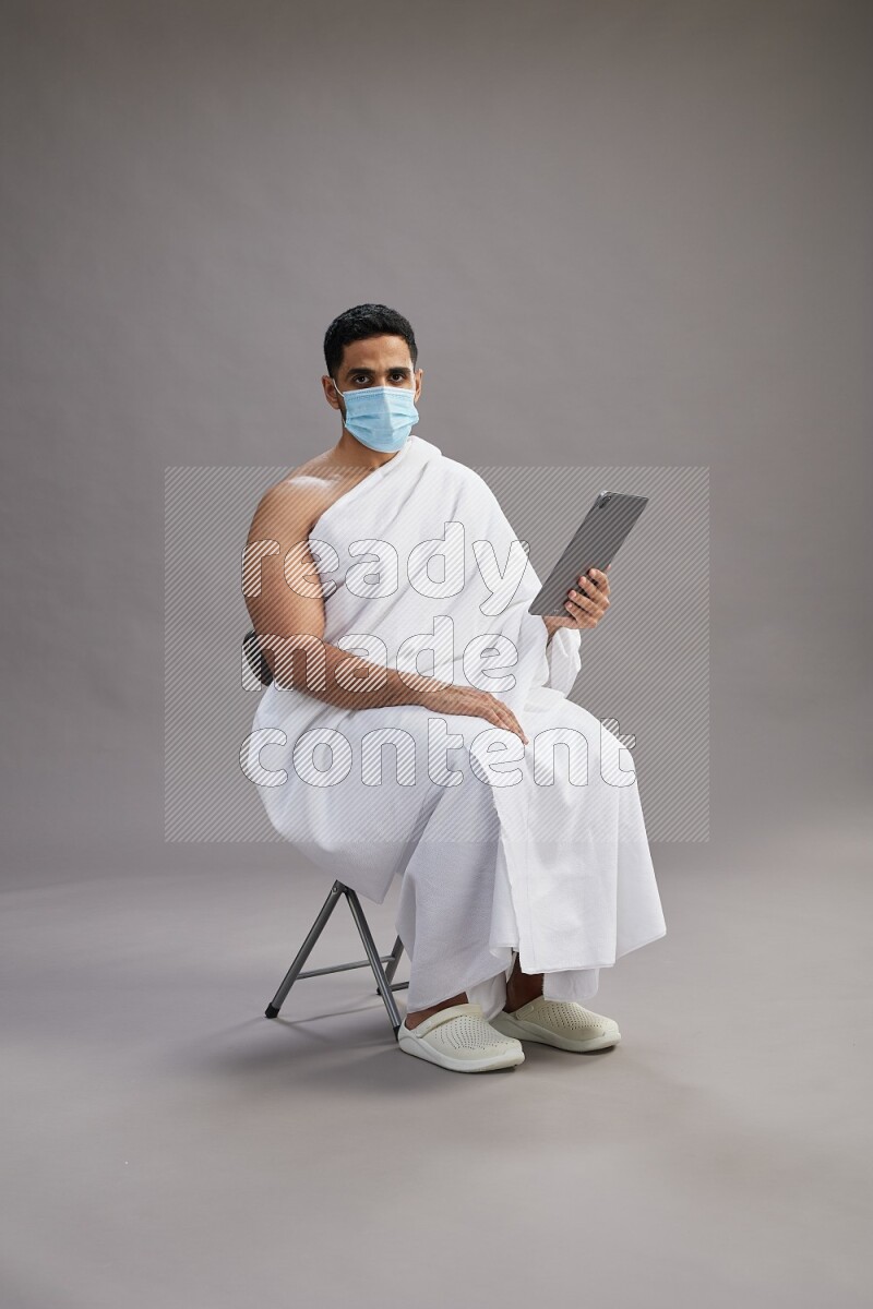 A man wearing Ehram with face mask sitting on chair holding a tablet on gray background