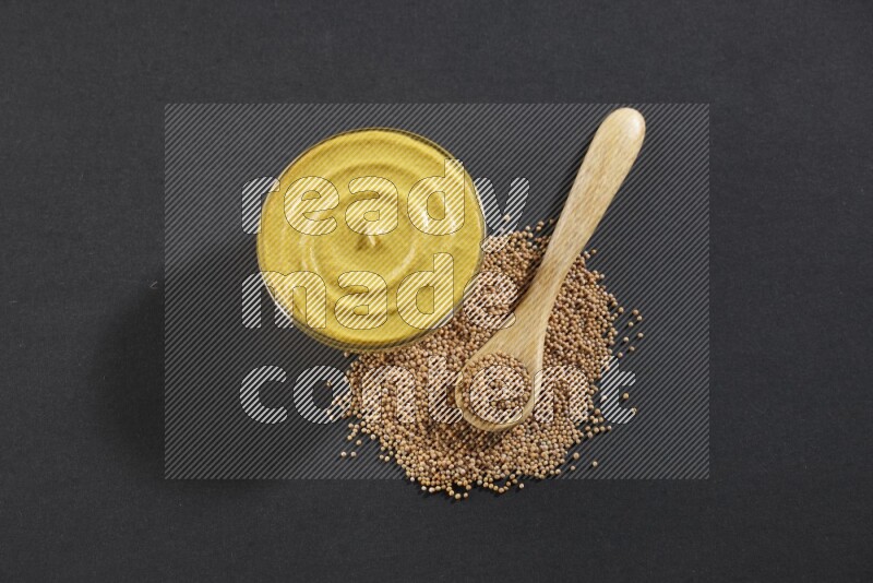 A glass bowl full of mustard paste with mustard seeds underneath and a full wooden spoon on black flooring