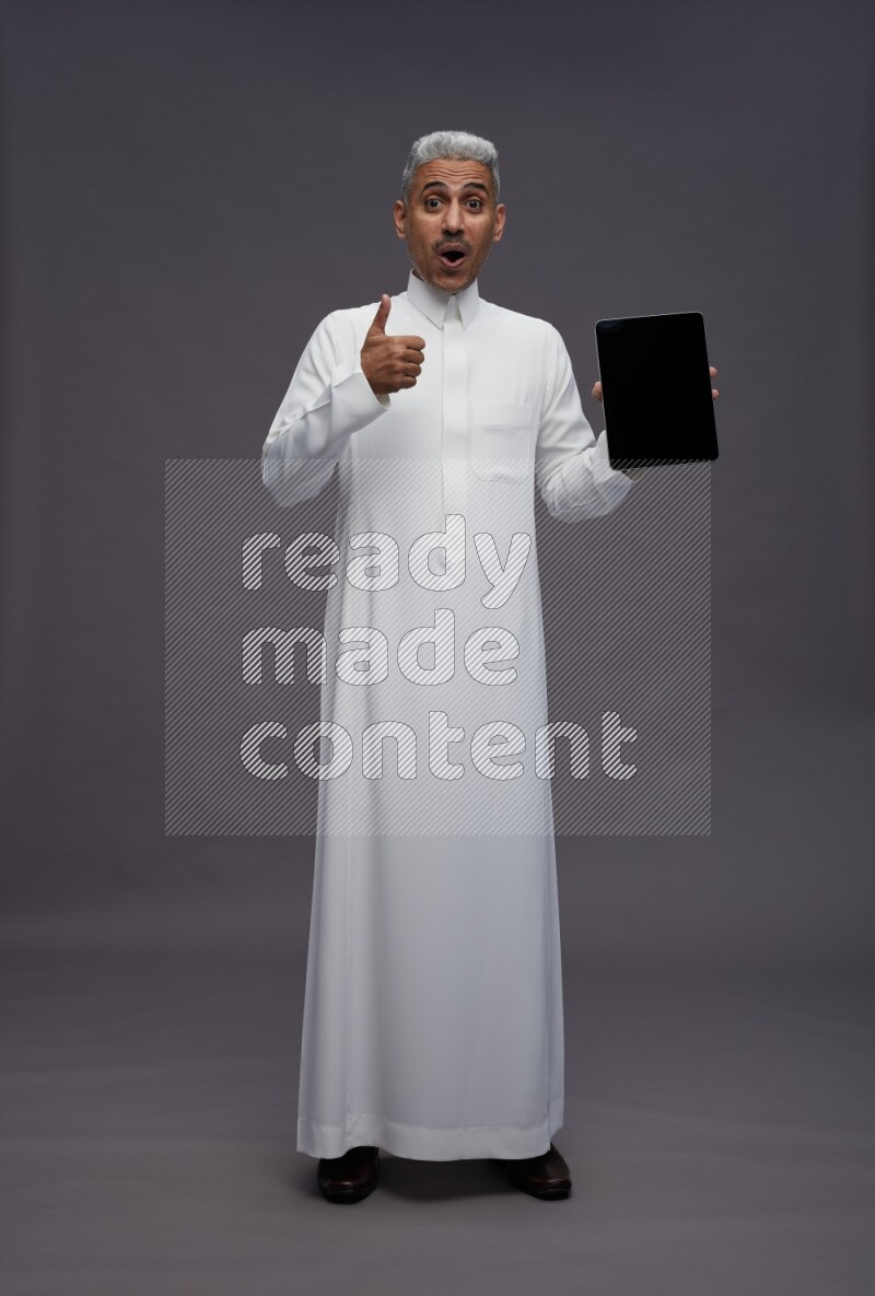 Saudi man wearing thob standing showing tablet to camera on gray background