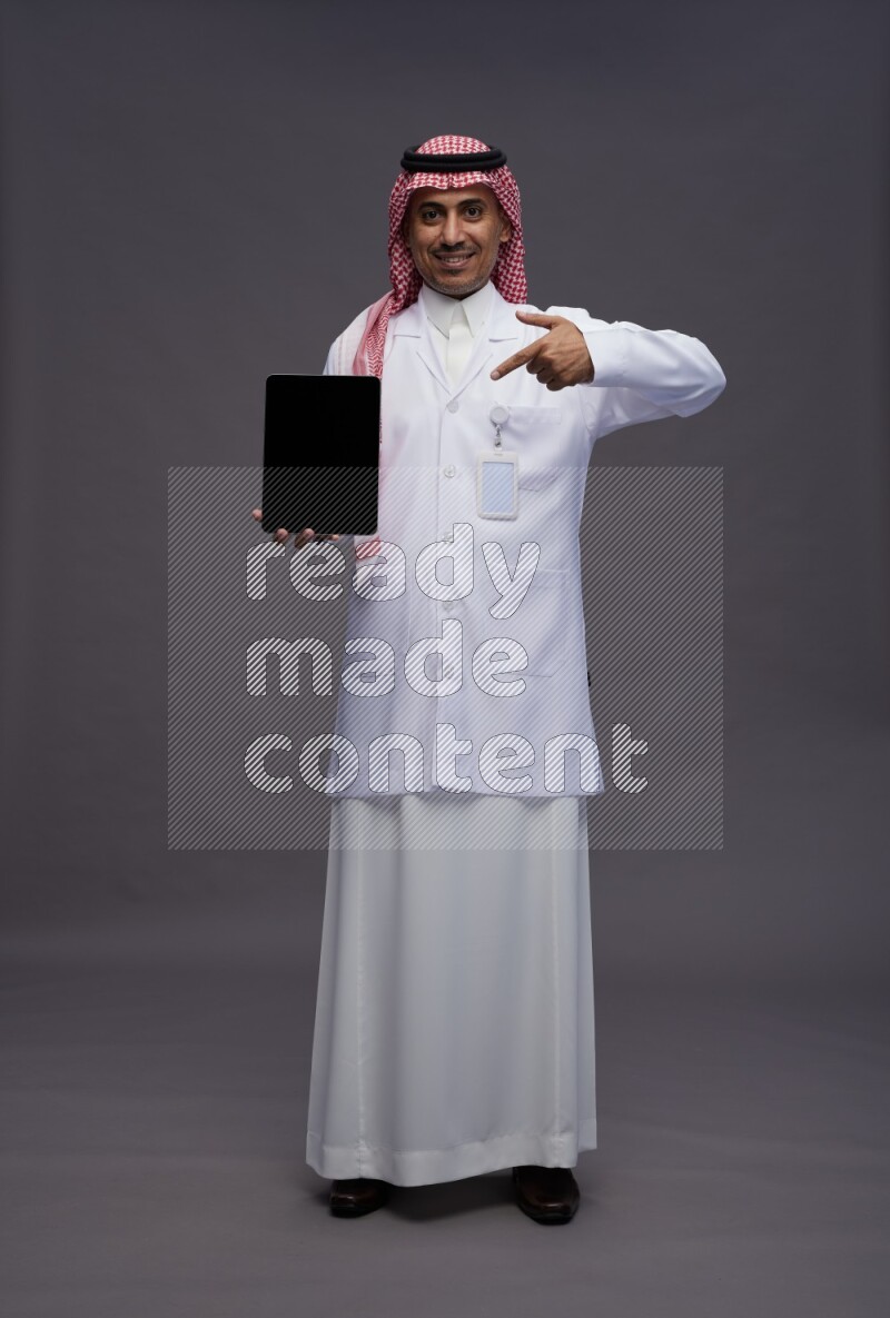 Saudi man wearing thob with lab coat and shomag with pocket employee badge standing showing tablet to camera on gray background