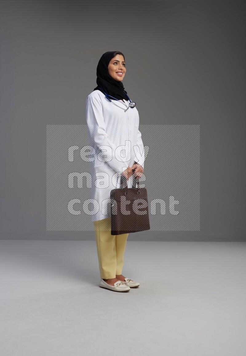 Saudi woman wearing lab coat with stethoscope standing holding bag on Gray background