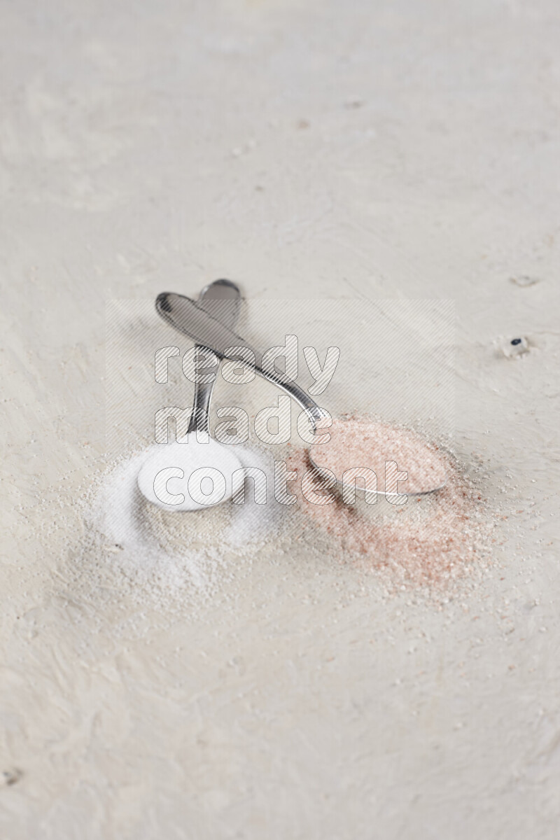 2 metal spoons filled with white salt and pink himalayan salt on white background