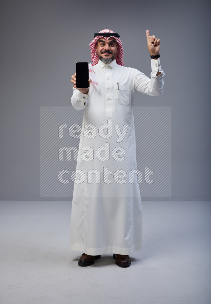 Saudi man Wearing Thob and red Shomag standing showing phone to camera on Gray background