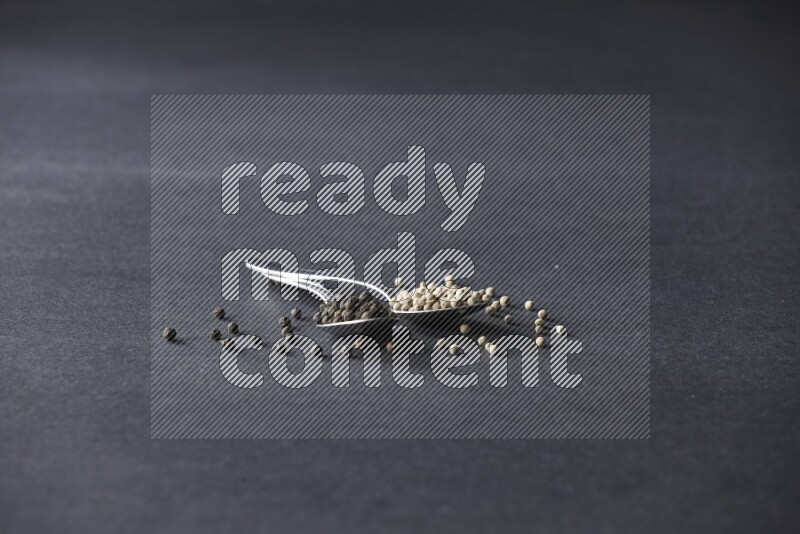 ملعقتان معدنيتان ممتلئتان بحبوب الفلفل الأسود والأبيض مع حبوب متناثرة على أرضية سوداء