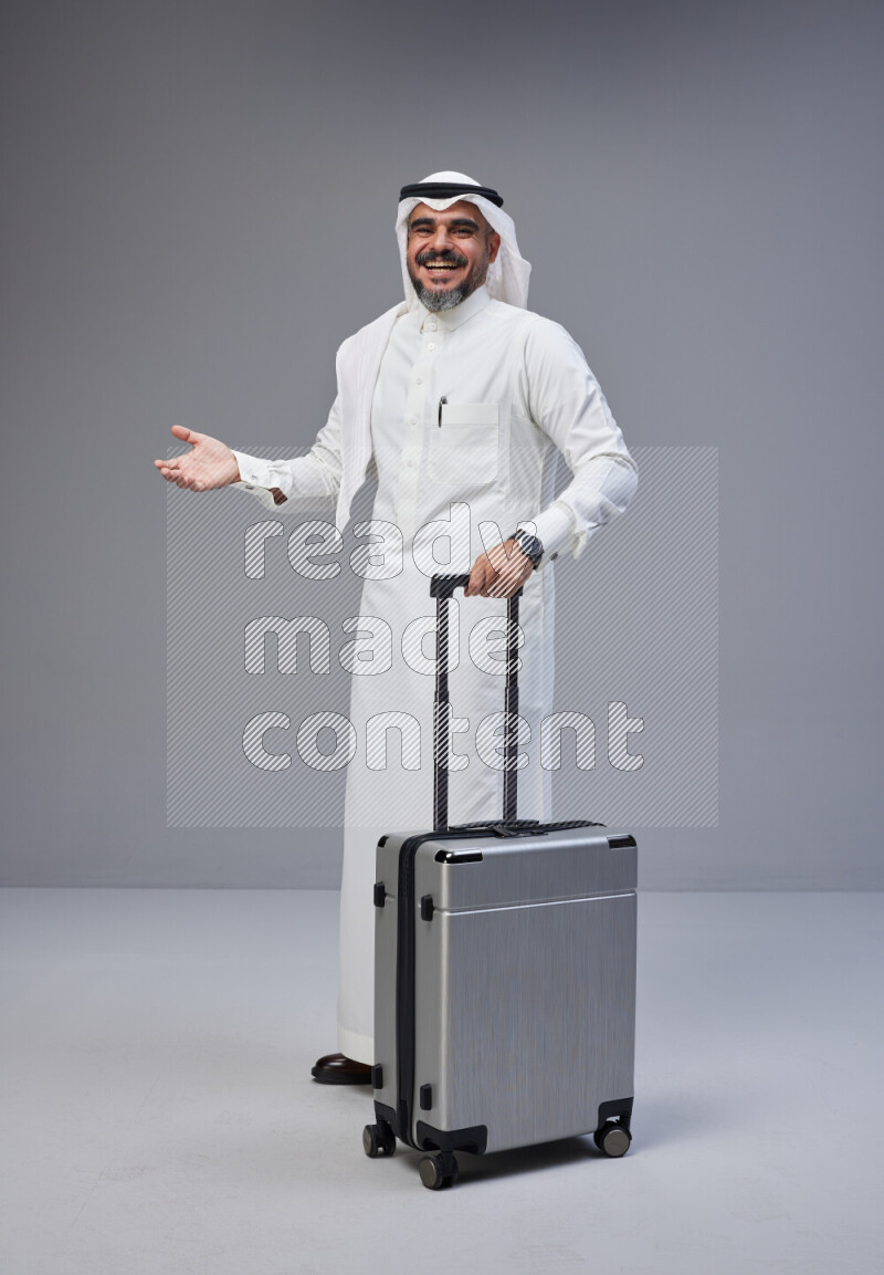 Saudi man wearing Thob and white Shomag standing holding Travel bag on Gray background