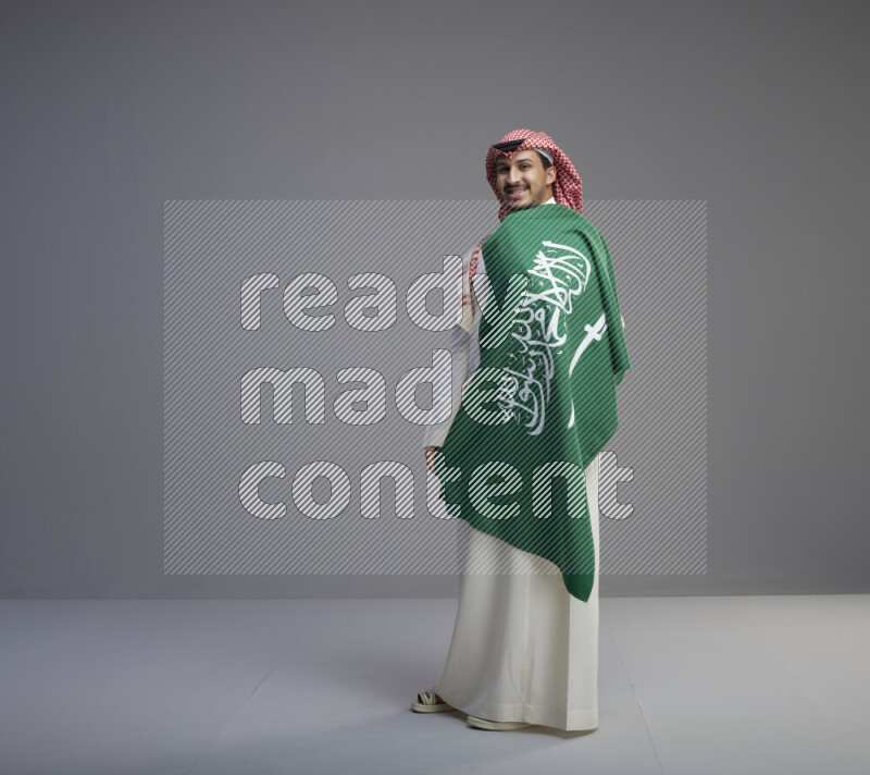 A saudi man standing wearing thob and red shomag holding big saudi flag on gray background