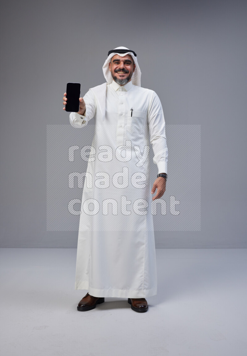 Saudi man Wearing Thob and white Shomag standing showing phone to camera on Gray background
