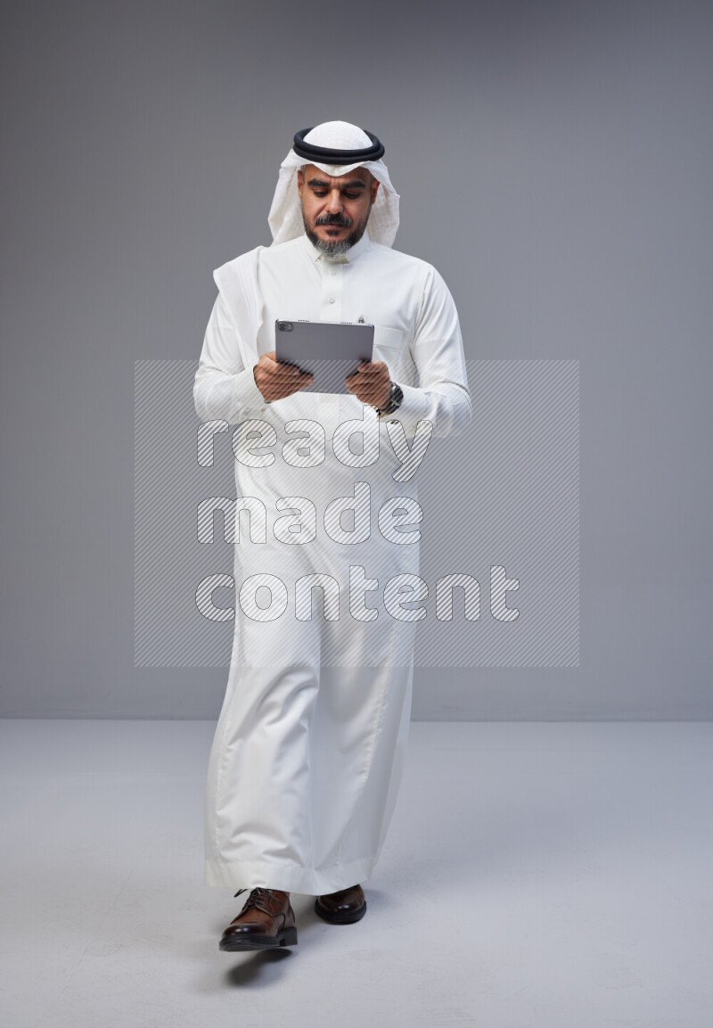 Saudi man Wearing Thob and white Shomag standing working on tablet on Gray background