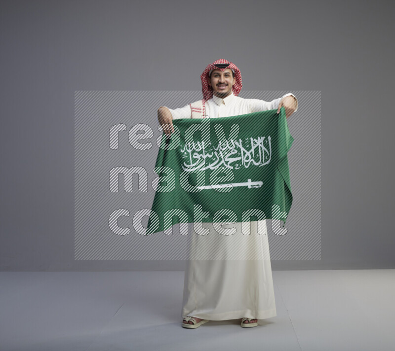 A saudi man standing wearing thob and red shomag holding big saudi flag on gray background