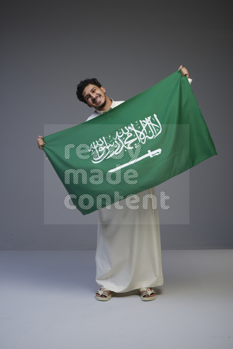 A Saudi man standing wearing thob holding big Saudi flag on gray background