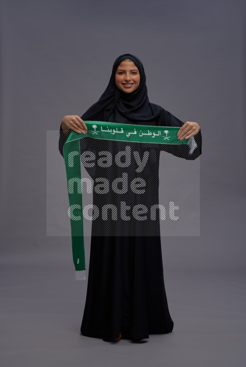 Saudi woman wearing Abaya standing holding Saudi flag on gray background