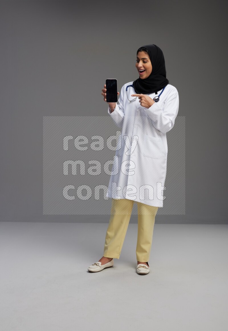 Saudi woman wearing lab coat with stethoscope standing showing phone to camera with sign in the back on Gray background