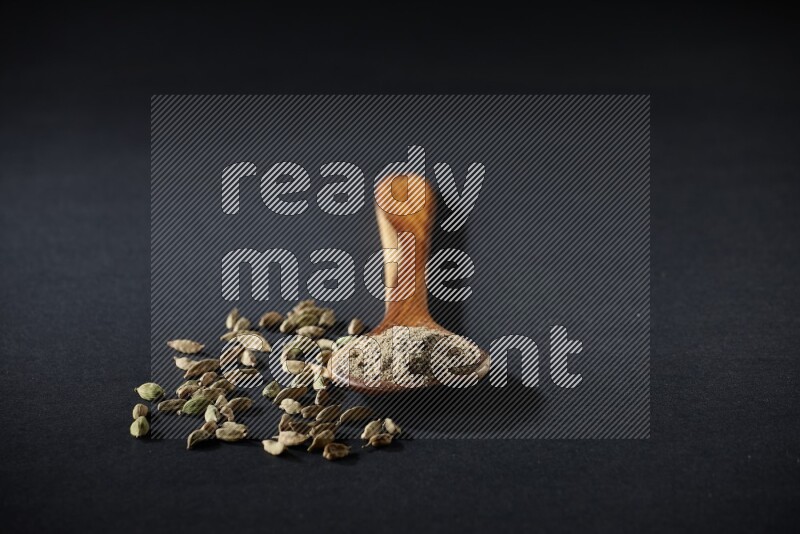 A wooden ladle full of cardamom powder and cardamom seeds beside it on black flooring