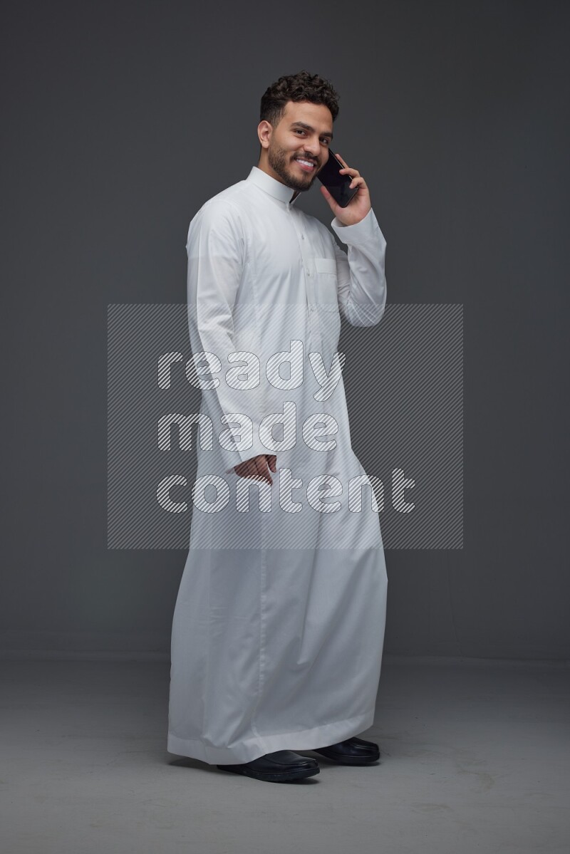 A Saudi man wearing Thobe and talking in the phone while standing and making different poses eye level on a gray background