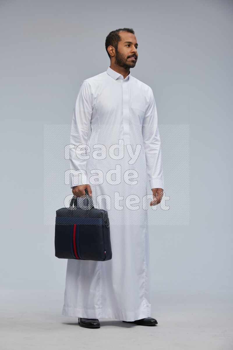 Saudi man wearing thob standing holding suitcase on gray background