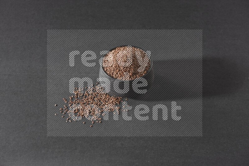 A black pottery bowl full of flaxseeds surrounded by the seeds on a black flooring