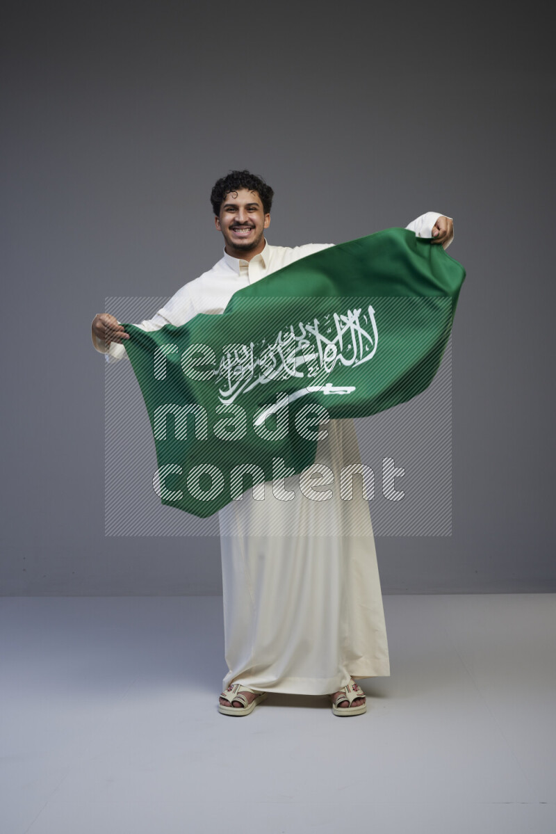 A Saudi man standing wearing thob holding big Saudi flag on gray background