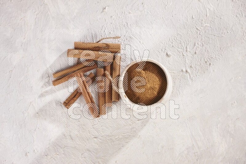 Cinnamon sticks stacked beside a beige bowl full of cinnamon powder on white background