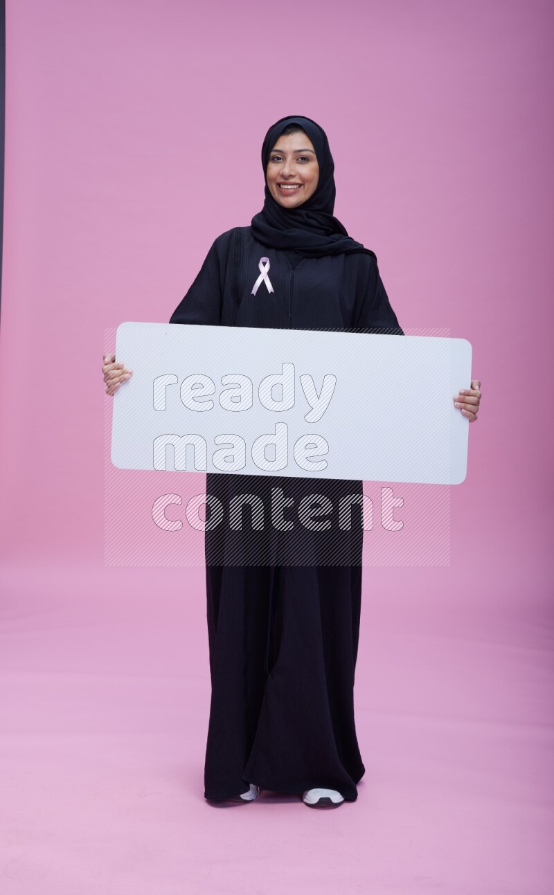 Saudi woman wearing pink ribbon on Abaya standing holding board on pink background