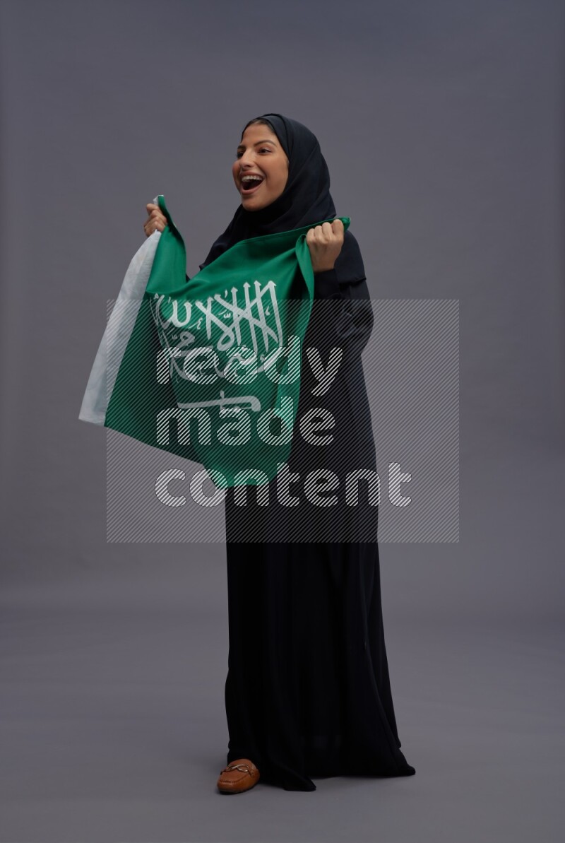 Saudi woman wearing Abaya standing holding Saudi flag on gray background
