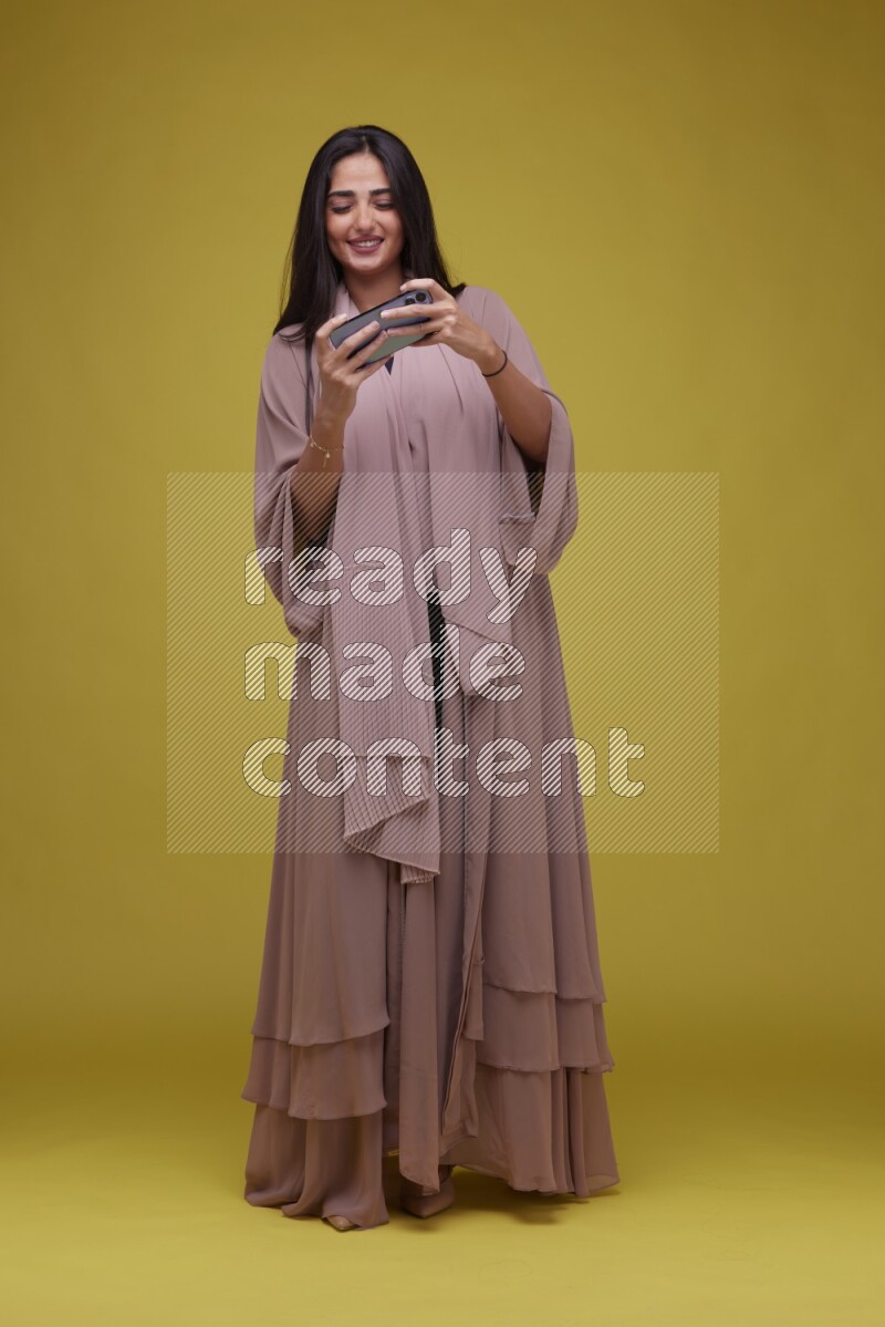 A woman Playing a Game on her smartphone on a Yellow Background wearing Brown Abaya
