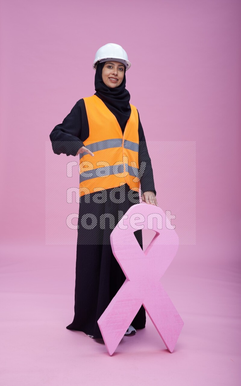 Saudi woman wearing Abaya with engineer vest and helmet standing awareness ribbon on pink background