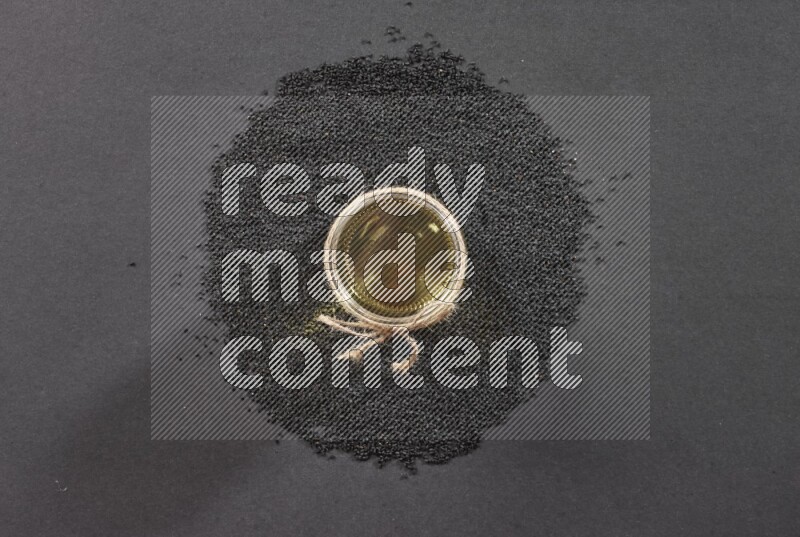 A glass jar full of black seeds oil surrounded by the seeds on a black flooring