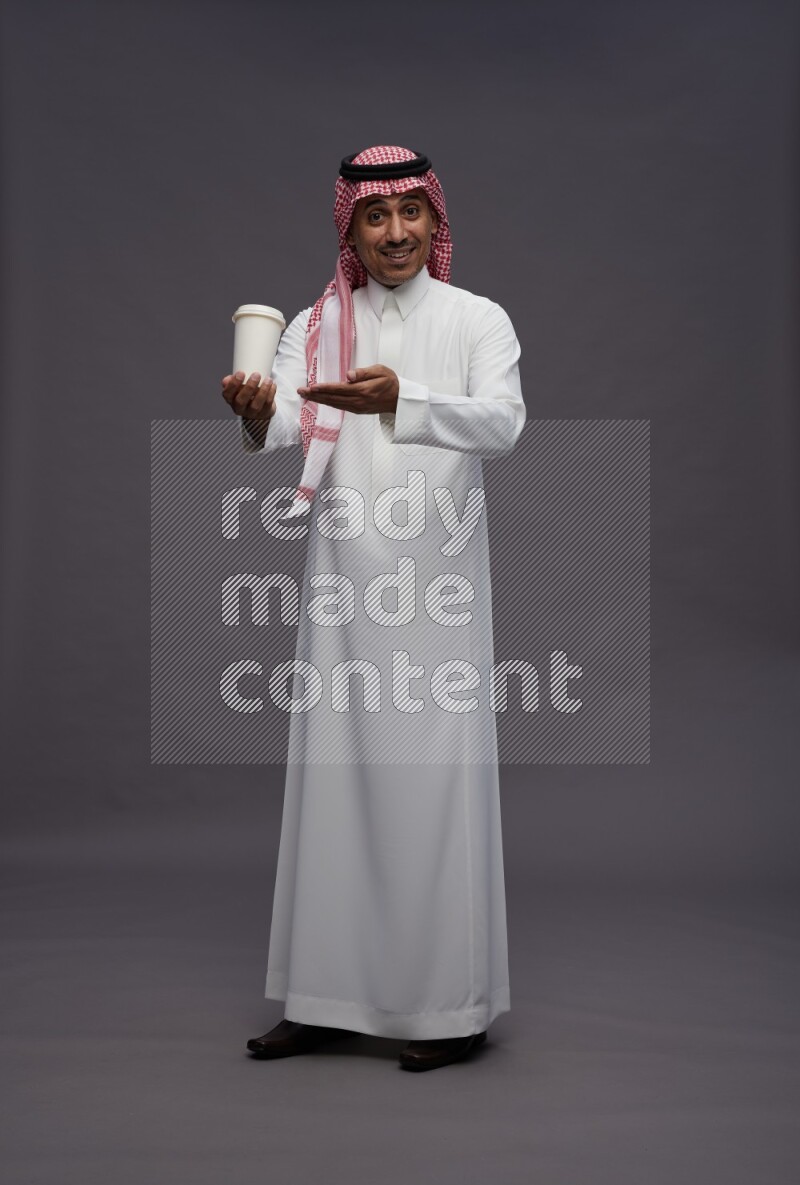 Saudi man wearing thob and shomag standing holding paper cup on gray background