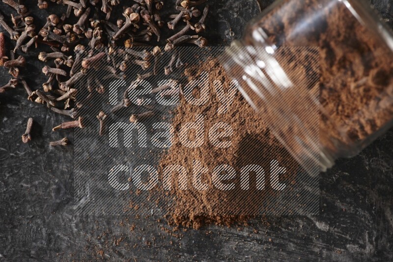 A flipped glass jar full of cloves powder with cloves spread on a textured black flooring