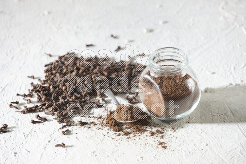 A glass spice jar and a metal spoon full of cloves powder and cloves spread on textured white flooring