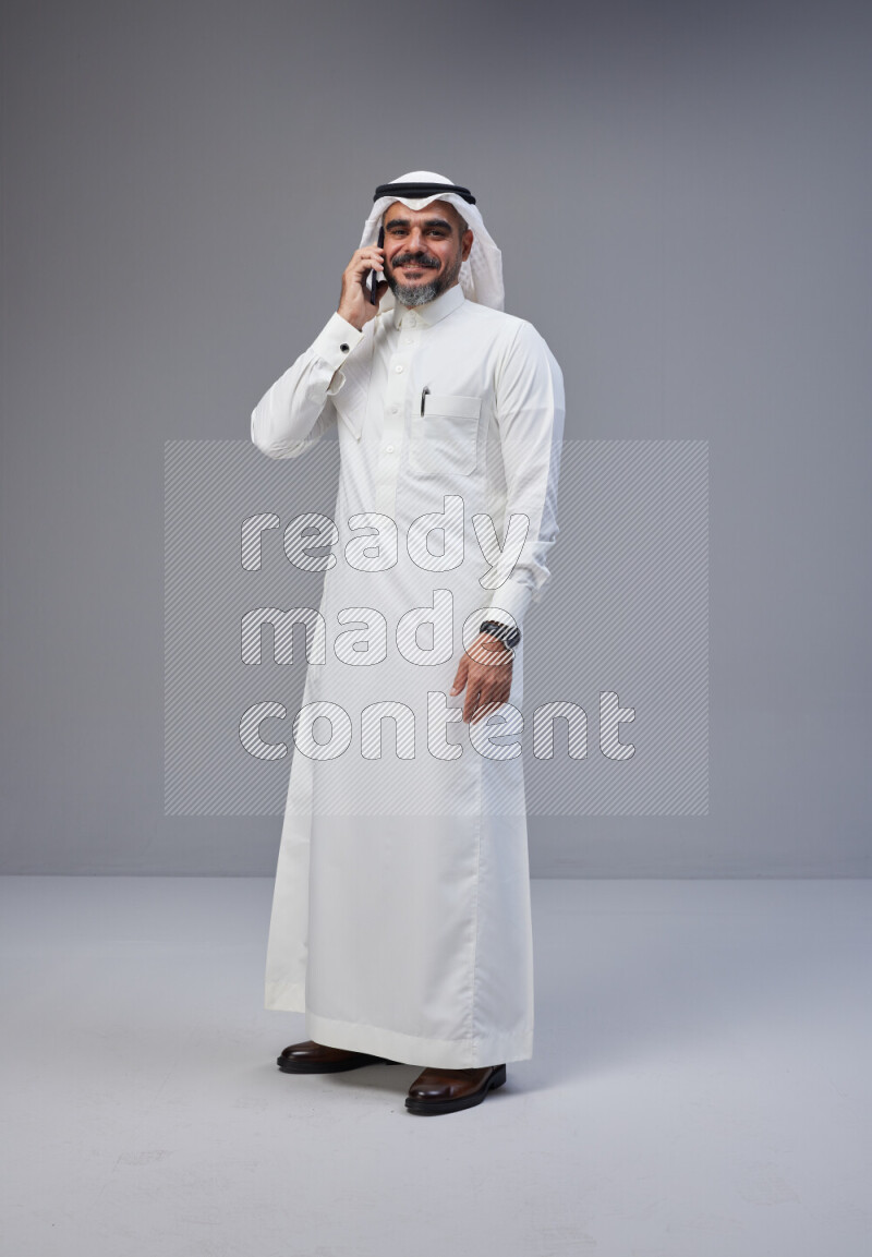 Saudi man Wearing Thob and white Shomag standing talking on phone on Gray background