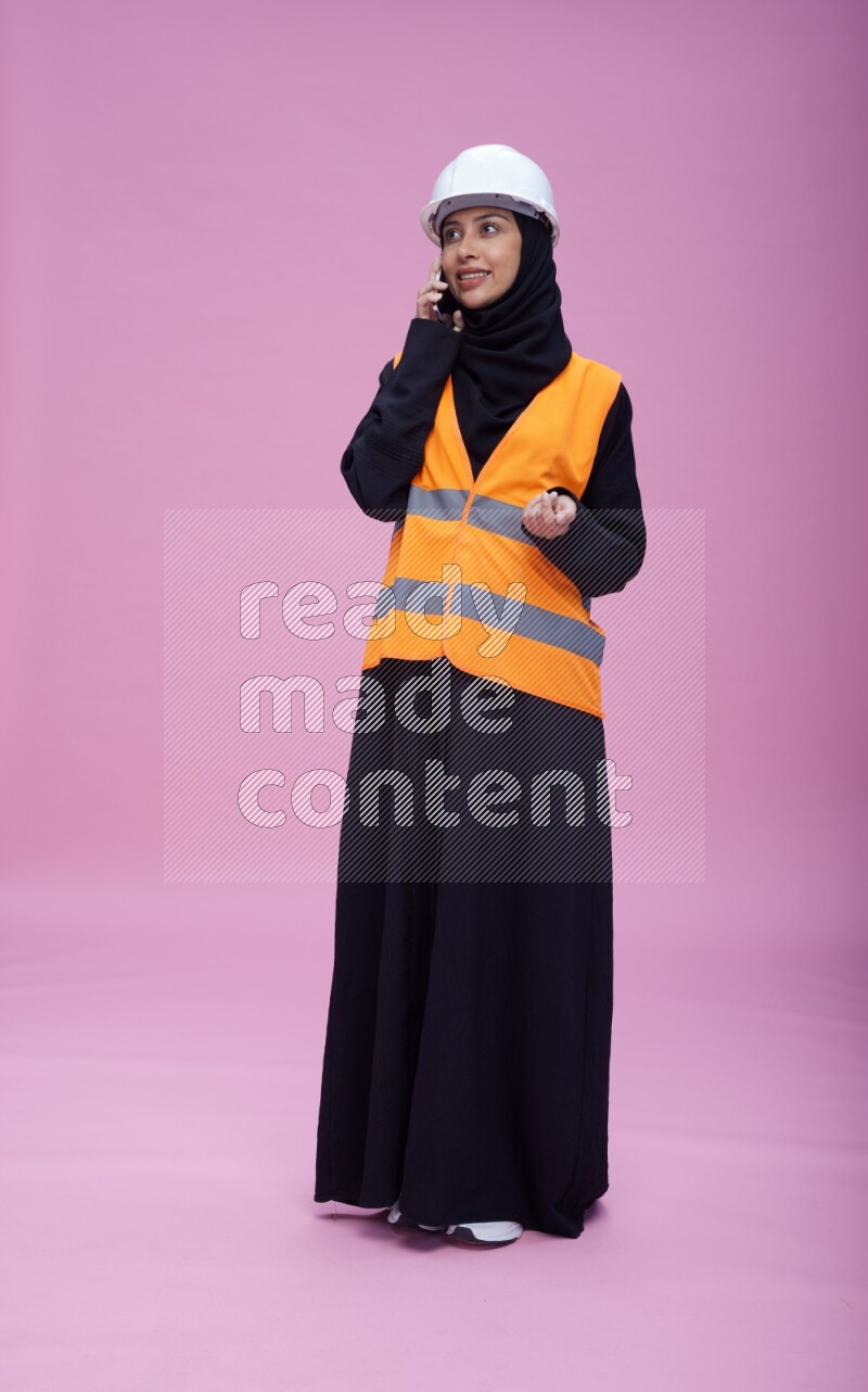Saudi woman wearing Abaya with engineer vest and helmet standing talking on phone on pink background