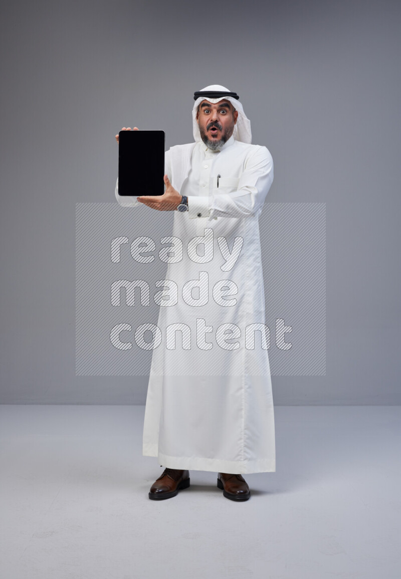 Saudi man Wearing Thob and white Shomag standing showing tablet to camera on Gray background