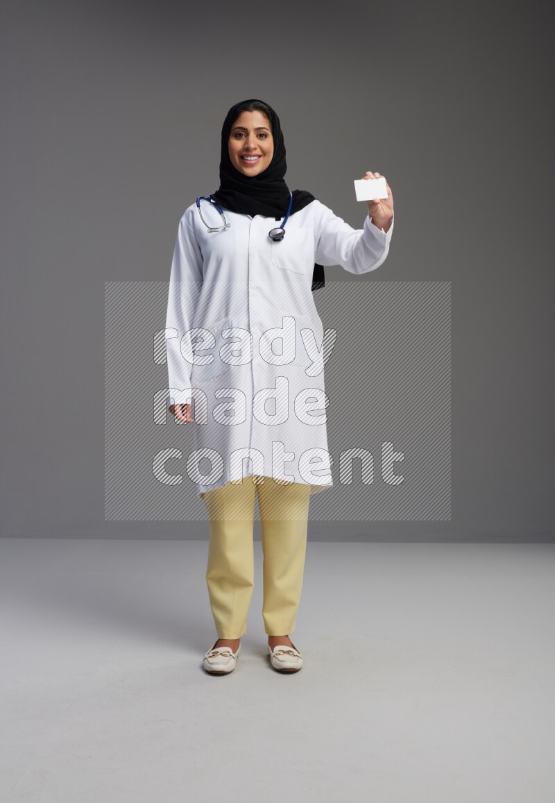 Saudi woman wearing lab coat with stethoscope standing holding ATM card on Gray background