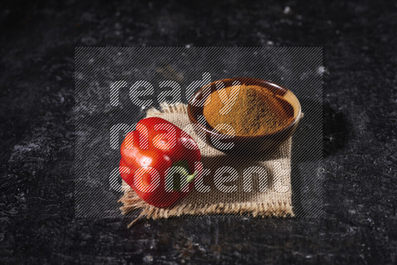 A wooden bowl full of ground paprika powder with a red bell pepper on a burlap fabric on black background