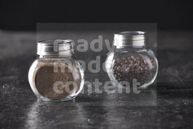 2 glass spice jars full of black pepper powder and black pepper beads on textured black flooring