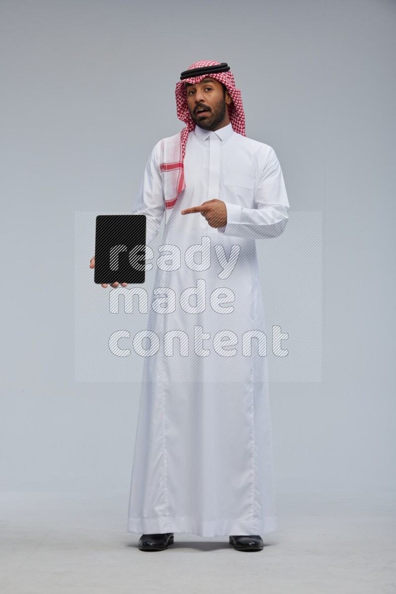 Saudi man Wearing Thob and shomag standing showing tablet to camera on Gray background