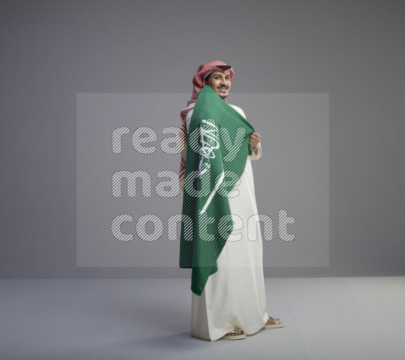A saudi man standing wearing thob and red shomag holding big saudi flag on gray background