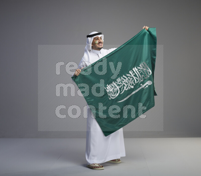 A Saudi man standing wearing thob and white shomag with face painting holding big Saudi flag on gray background