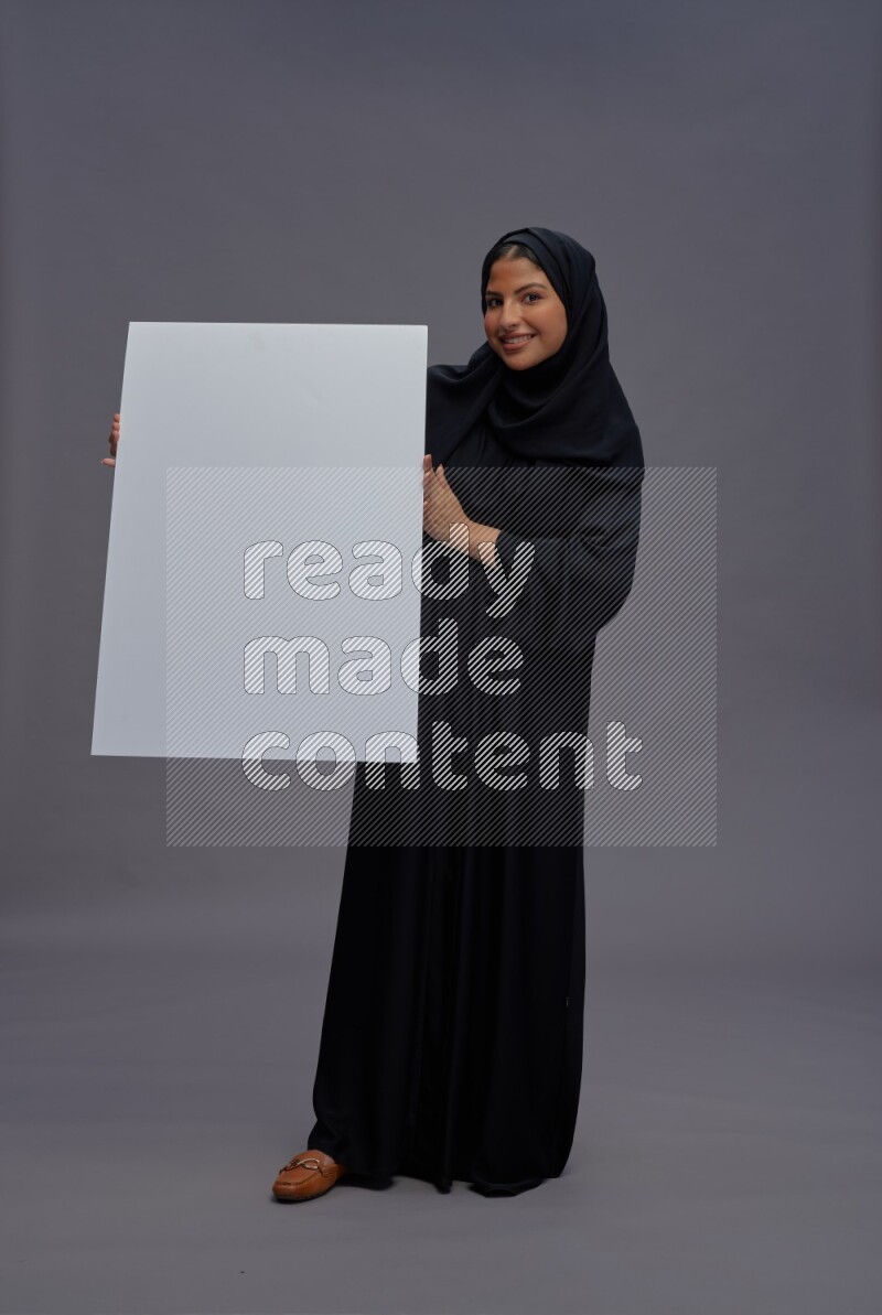 Saudi woman wearing Abaya standing holding white board on gray background