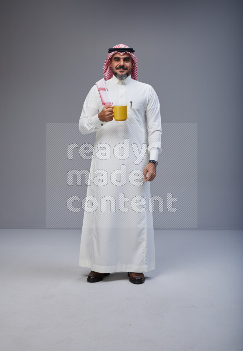 Saudi man Wearing Thob and red Shomag standing holding mug on Gray background