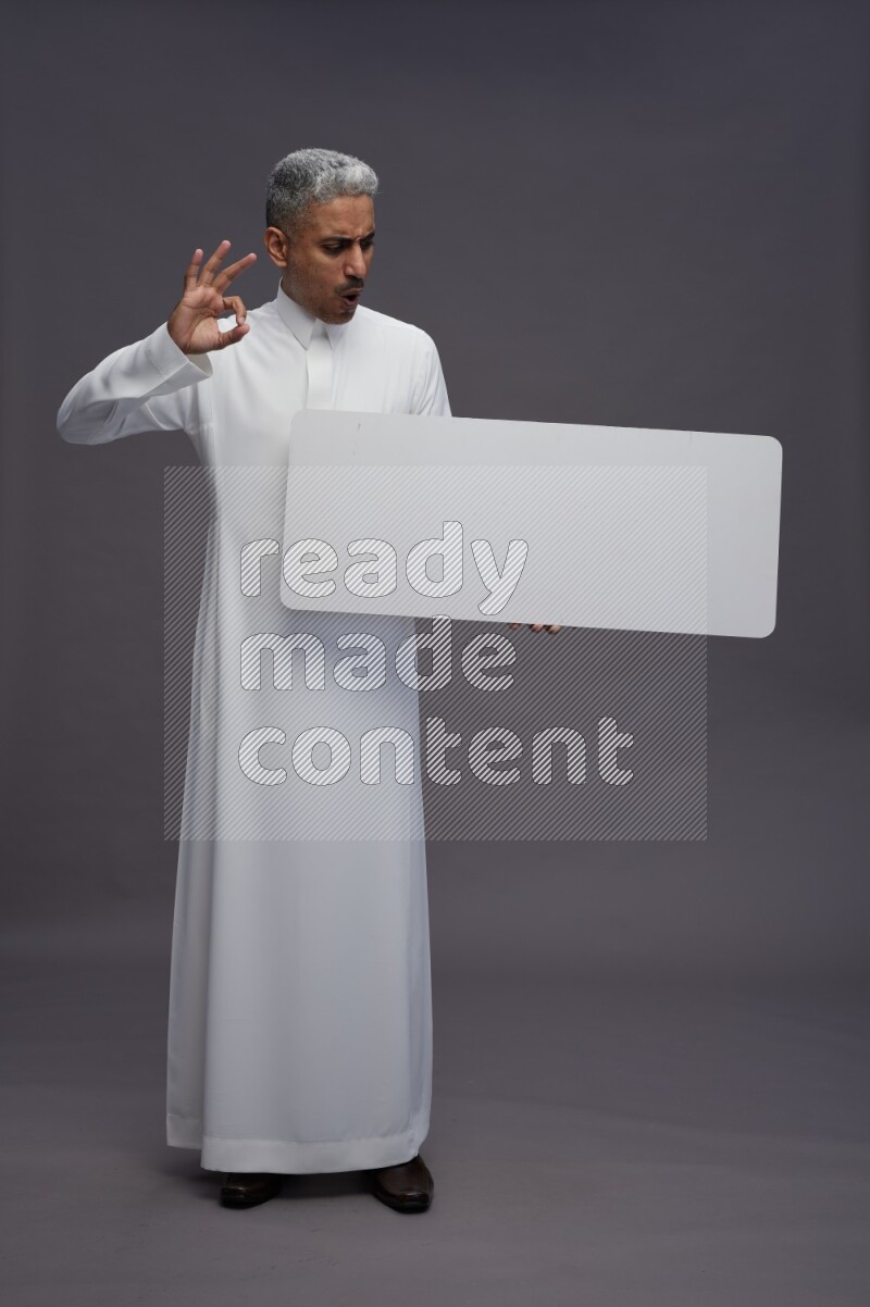 Saudi man wearing thob standing holding board on gray background
