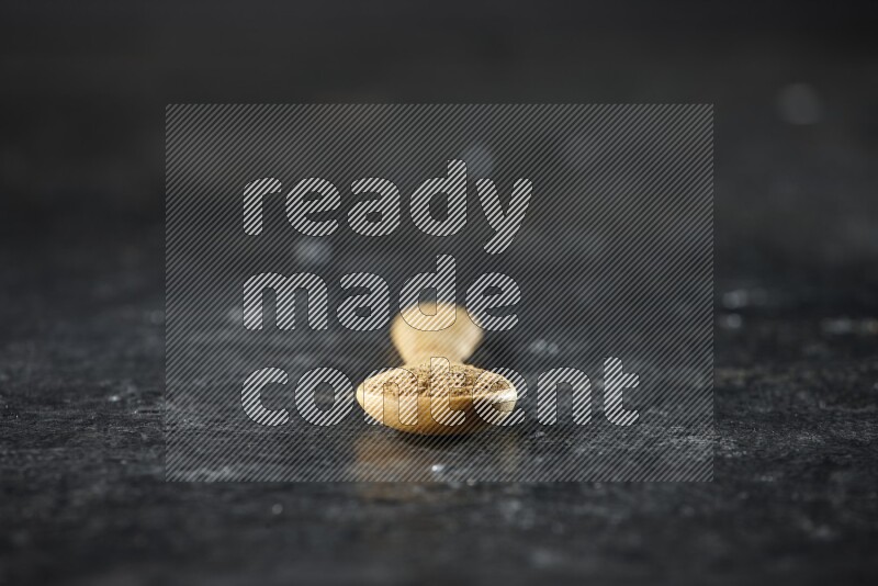A wooden spoons full of cumin powder on textured black flooring