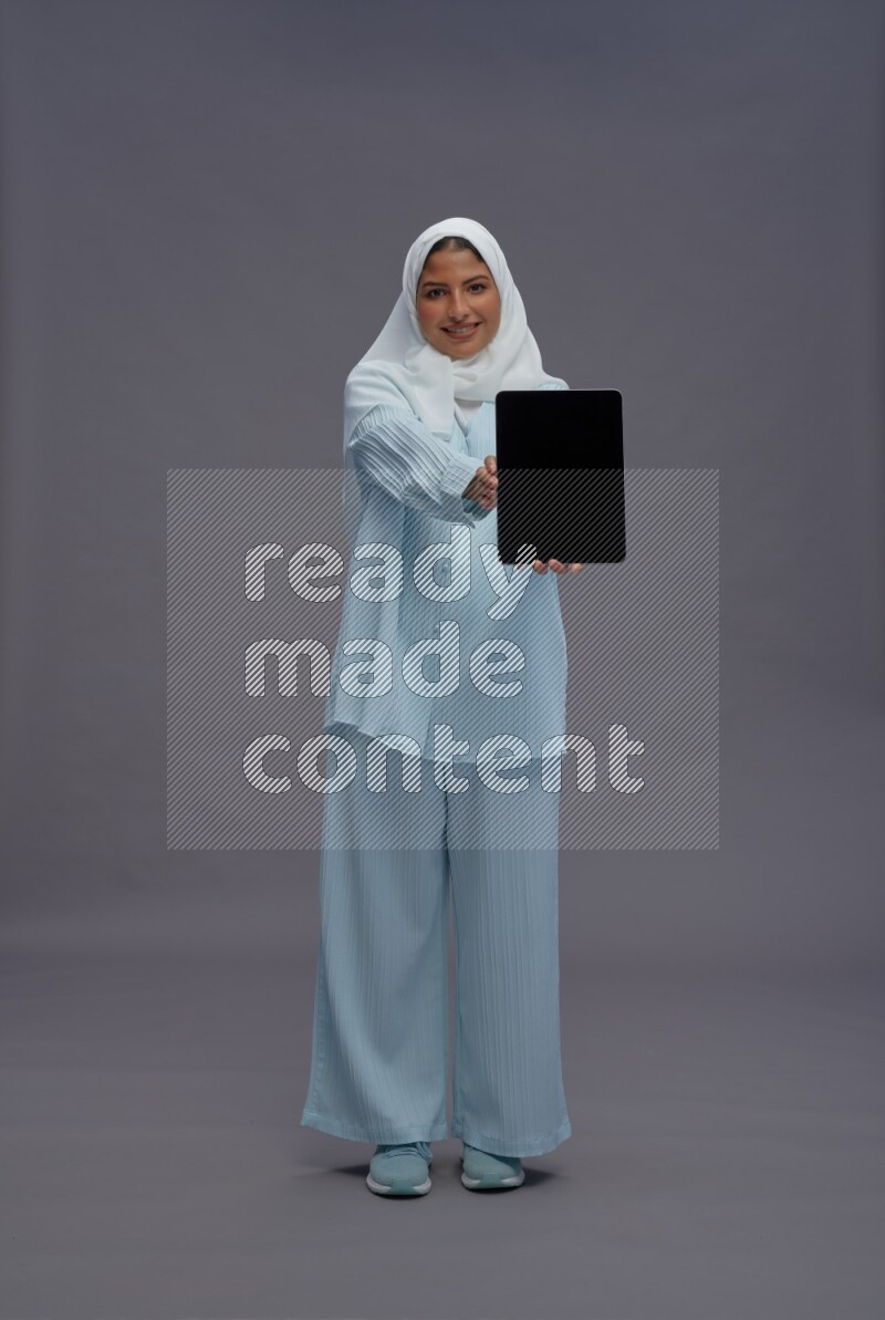 Saudi woman wearing hijab clothes standing showing tablet to camera on gray background
