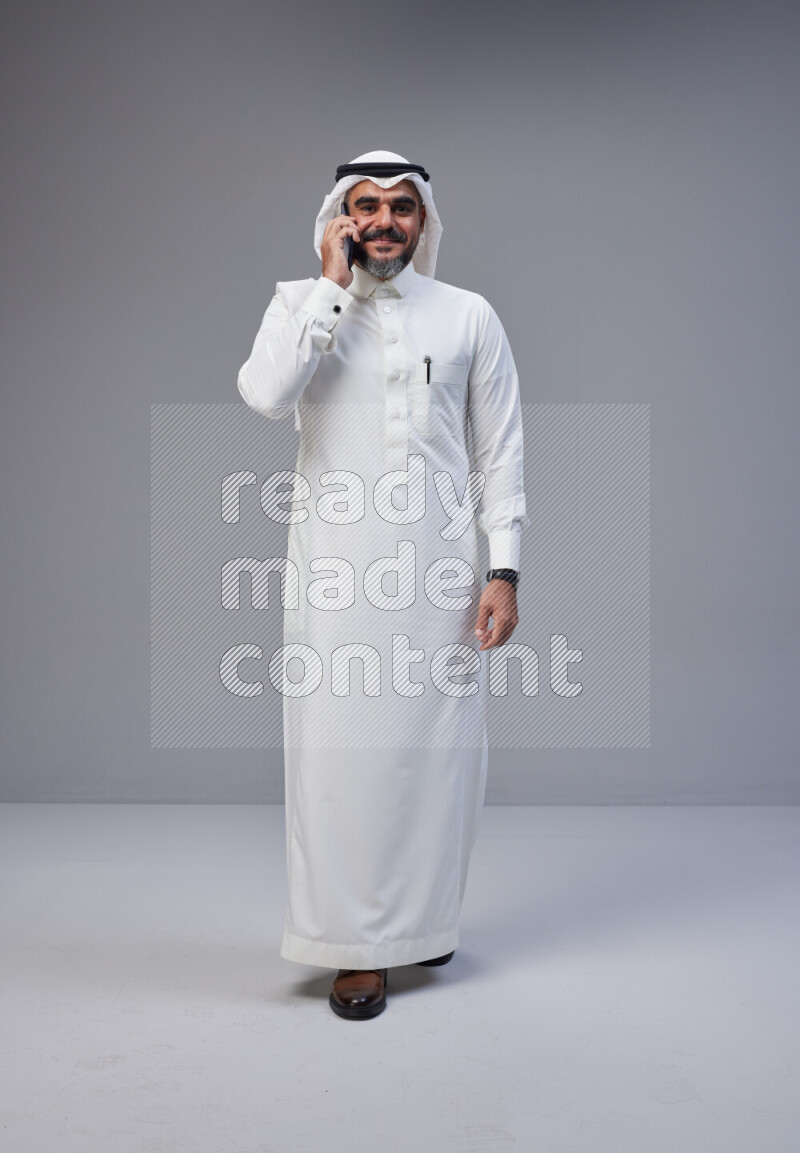 Saudi man Wearing Thob and white Shomag standing talking on phone on Gray background
