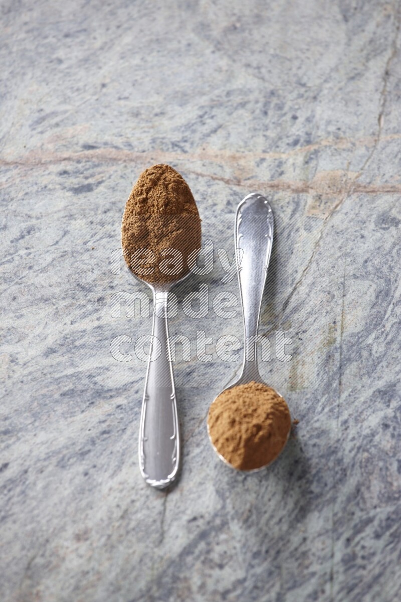 Two metal spoons full of cinnamon powder on grey marble background