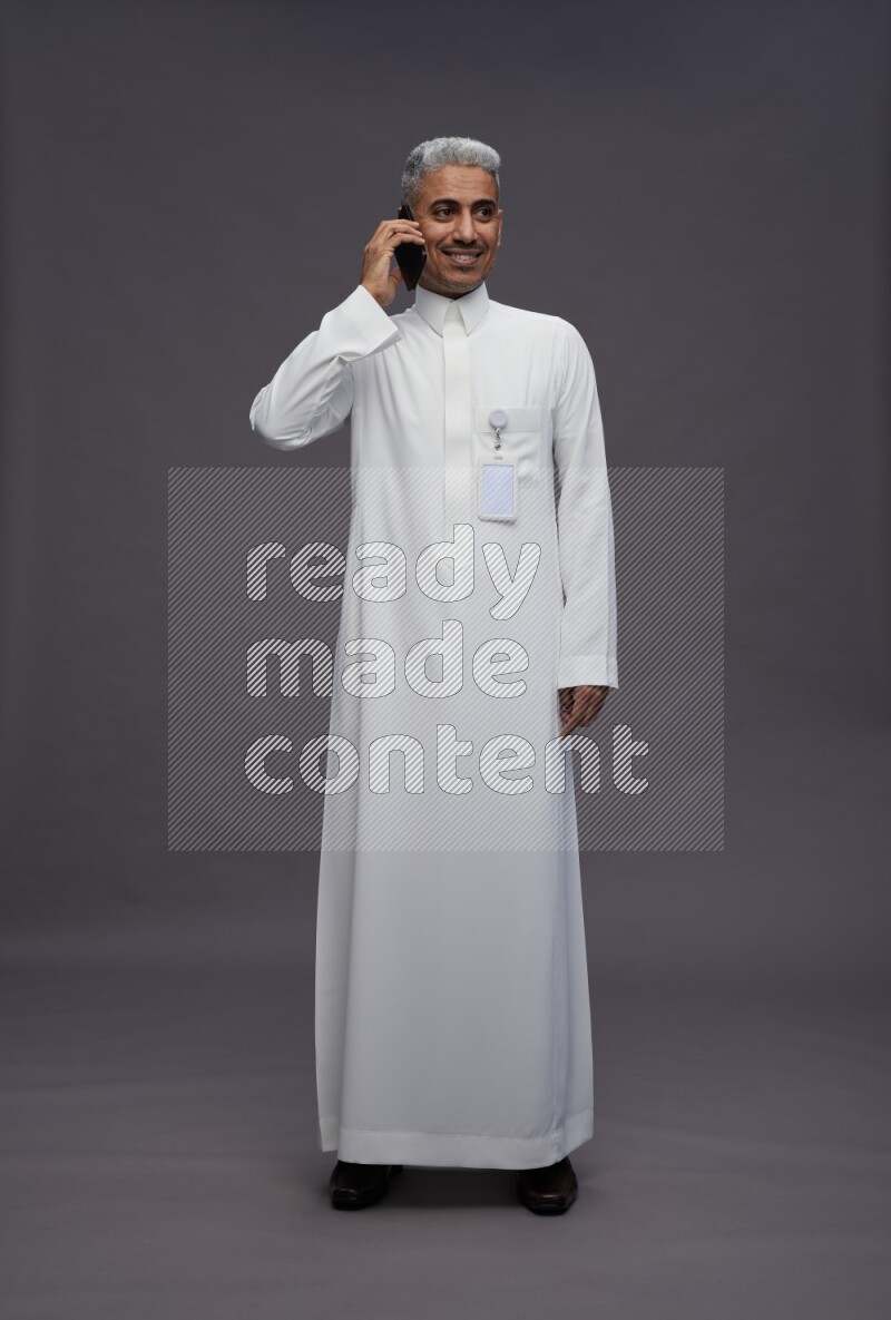 Saudi man wearing thob with pocket employee badge standing talking on phone on gray background