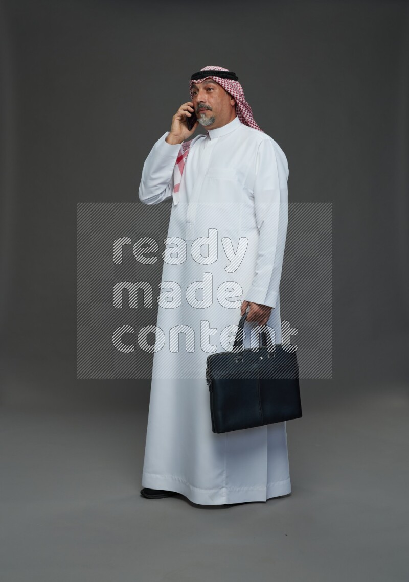 Saudi man with shomag Standing holding bag talking on phone on gray background