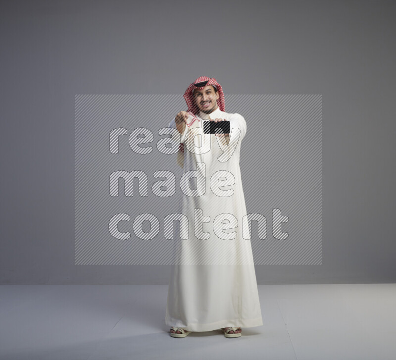 A Saudi man standing wearing thob and red shomag showing phone to camera on gray background